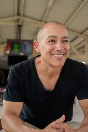 Portrait of a mature bald Hispanic man tourist in a black t-shirt sitting at a train station...