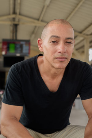 Portrait of a mature bald Hispanic man tourist in a black t-shirt sitting at a train station...