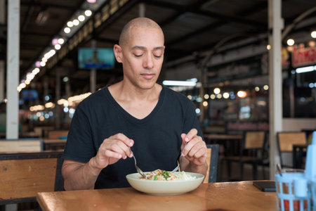 Portrait of a middle-aged bald Hispanic tourist man enjoying traditional Thai pad Thai noodles at an outdoor food court restaurant, smiling while dining and experiencing local cuisine.の写真素材