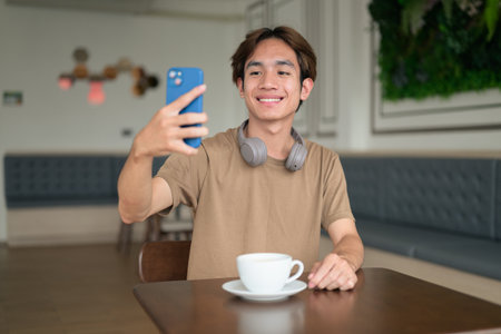 Young Thai student man in a brown t-shirt in coffee shop wearing headphones around his neck in a modern cafe concept of social media, remote communication and digital lifestyle.の写真素材