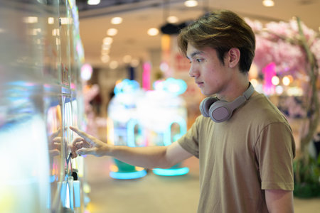 Young Thai Asian man in a brown t-shirt, with headphones around his neck, using a vending arcade...
