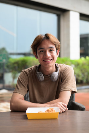 Young Asian man in brown t-shirt with headphones sitting at an outdoor tableの写真素材