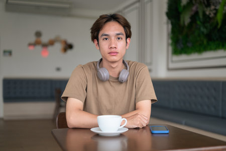 Young Thai student man in a brown t-shirt in coffee shop wearing headphones around his neck in a modern cafe concept of social media, remote communication and digital lifestyle.の写真素材