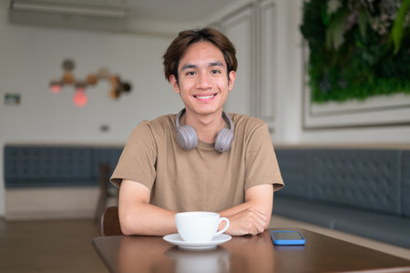 Young Thai student man in a brown t-shirt in coffee shop wearing headphones around his neck in a modern cafe concept of social media, remote communication and digital lifestyle.の写真素材