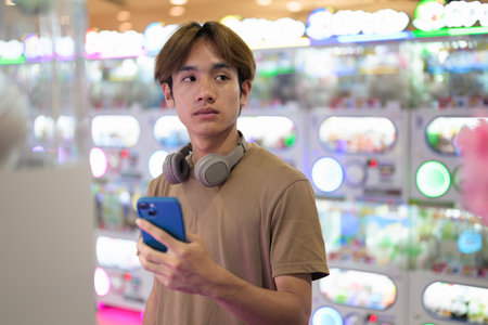 Young Thai Asian man in a brown t-shirt, with headphones around his neck, using a vending arcade machine and phone in a bright shopping mall, modern lifestyle, technology and leisure concept.の写真素材