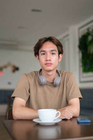 Young Thai student man in a brown t-shirt in coffee shop wearing headphones around his neck in a modern cafe concept of social media, remote communication and digital lifestyle.の写真素材