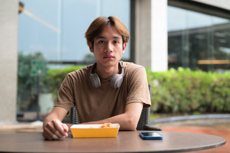 Young Asian student man in brown t-shirt with headphones ready to eat fried chicken drumstick at an outdoor table for fast food, snack and leisure meal conceptの写真素材