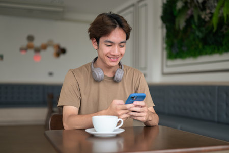 Young Thai student man in a brown t-shirt in coffee shop wearing headphones around his neck in a modern cafe concept of social media, remote communication and digital lifestyle.の写真素材