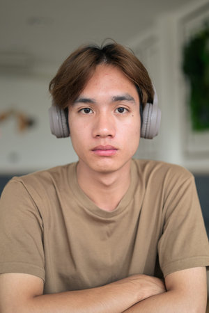Young Thai student man in a brown t-shirt in coffee shop wearing headphones around his neck in a...