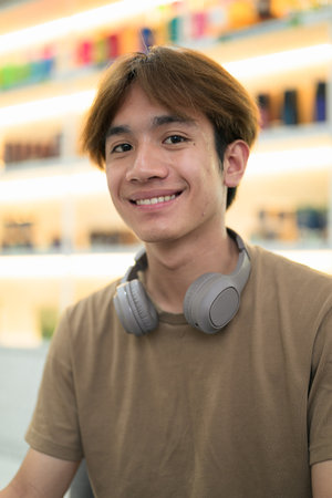 Young Thai student man in a brown t-shirt in coffee shop wearing headphones around his neck in a modern cafe concept of social media, remote communication and digital lifestyle.の写真素材