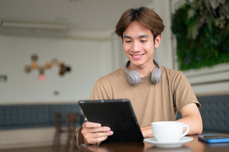 Young Thai student man in a brown t-shirt in coffee shop wearing headphones around his neck in a modern cafe concept of social media, remote communication and digital lifestyle.の写真素材