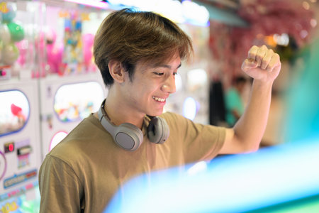 Young Thai Asian man in a brown t-shirt, with headphones around his neck, using a vending arcade machine in a bright shopping mall modern lifestyle, technology and leisure conceptの写真素材