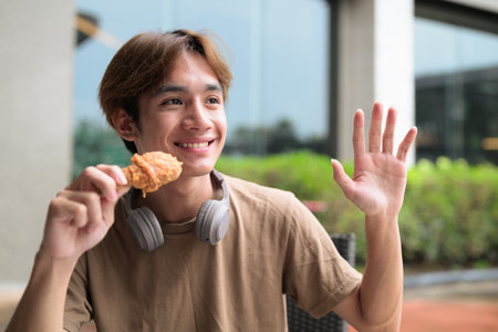 Young Asian student man in brown t-shirt with headphones ready to eat fried chicken drumstick at an outdoor table for fast food, snack and leisure meal conceptの写真素材