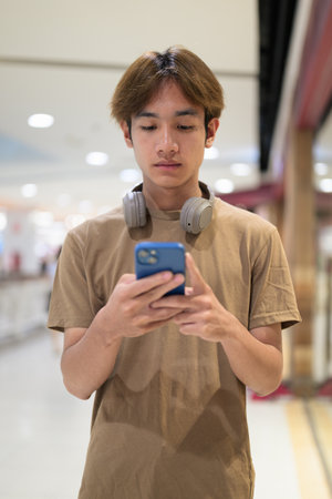 20-year-old Thai man with headphones around his neck looks down smiling at his phone in a bright mall corridor, messaging, online shopping and urban lifestyle.の写真素材
