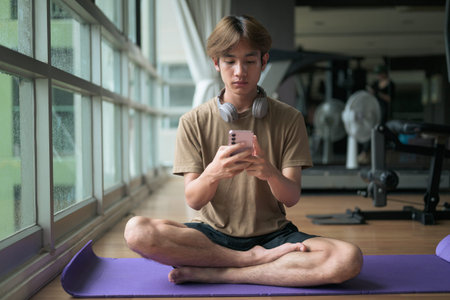 Young Thai Asian man with headphones sits cross-legged on a yoga mat near a large window in a...