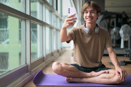 Young Thai Asian man with headphones sits cross-legged on a yoga mat near a large window in a fitness studio, focused on his smartphone with wellness, post-workout and mindfulness with technology.の写真素材