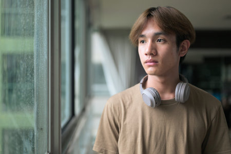 Young handsome and slim Asian Thai man with headphones next to window in gym