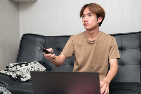 Young Thai Asian man wearing a brown t-shirt, sitting on a black leather sofa while using a laptop at home concept of modern lifestyle, remote work, study and digital technologyの写真素材