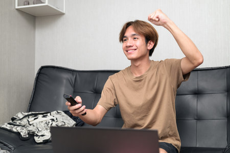 Young Thai Asian man wearing a brown t-shirt, sitting on a black leather sofa while using a laptop at home concept of modern lifestyle, remote work, study and digital technologyの写真素材