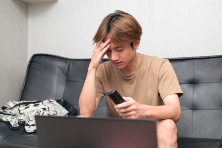 Young Thai Asian man wearing a brown t-shirt, sitting on a black leather sofa while using a laptop at home concept of modern lifestyle, remote work, study and digital technologyの写真素材