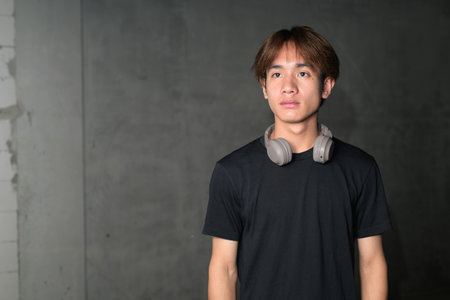 Young Thai Asian man wearing a black t-shirt, posing in a minimalist brutalist-style construction area with gray concrete walls concept of modern lifestyle, urban fashion and architectureの写真素材