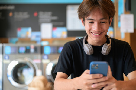 Young handsome Thai man sitting in a modern launderette with washing machines using phone, wearing a black t-shirt. Candid portrait showing Gen Z lifestyle, daily chores, laundry, and urban living.の写真素材