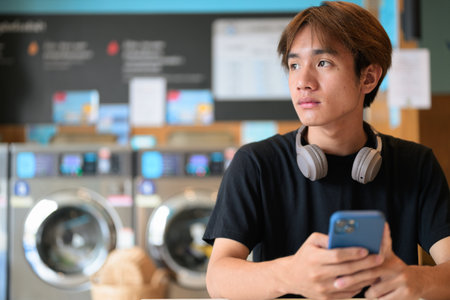 Young handsome Thai man sitting in a modern launderette with washing machines using phone, wearing a black t-shirt. Candid portrait showing Gen Z lifestyle, daily chores, laundry, and urban living.の写真素材