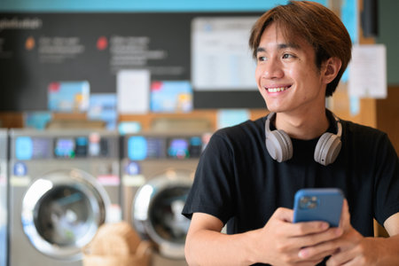 Young handsome Thai man sitting in a modern launderette with washing machines using phone, wearing a black t-shirt. Candid portrait showing Gen Z lifestyle, daily chores, laundry, and urban living.の写真素材