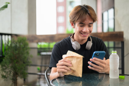Happy young Asian Thai student man eating sandwich outdoors for casual lunch while sitting and using phoneの写真素材