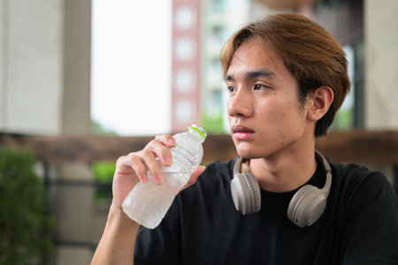 Young man drinking water from bottle while sitting outdoors and having headphones around his neckの写真素材