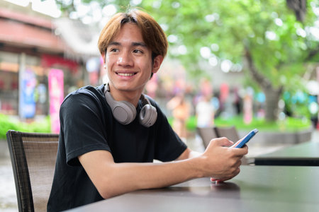 Portrait of a young Thai Asian man in casual style sitting and using phone outdoors during summer. Confident, relaxed, and authentic look suitable for various concepts and usesの写真素材