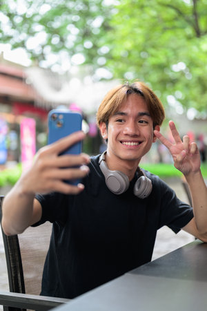 Portrait of a young Thai Asian man in casual style sitting and using phone outdoors during summer. Confident, relaxed, and authentic look suitable for various concepts and usesの写真素材