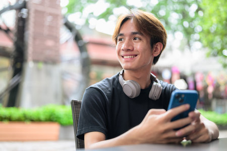 Portrait of a young Thai Asian man in casual style sitting and using phone outdoors during summer. Confident, relaxed, and authentic look suitable for various concepts and usesの写真素材