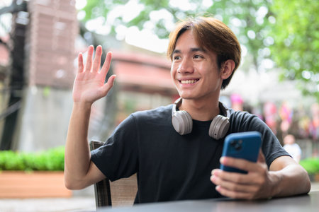 Portrait of a young Thai Asian man in casual style sitting and using phone outdoors during summer. Confident, relaxed, and authentic look suitable for various concepts and usesの写真素材