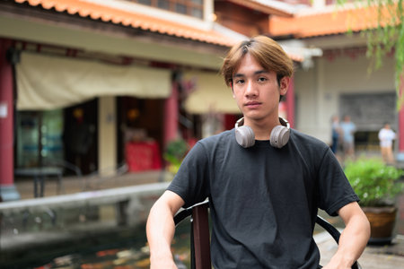 Young Thai Asian man sitting outdoors in traditional Chinese garden market. Confident, relaxed, and authentic look suitable for various concepts and usesの写真素材