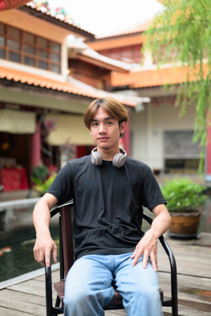 Young Thai Asian man sitting outdoors in traditional Chinese garden market. Confident, relaxed, and authentic look suitable for various concepts and usesの写真素材