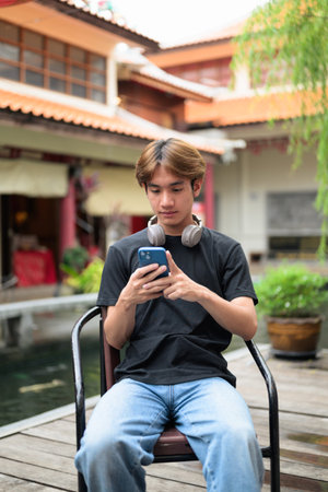 Young Thai Asian man sitting outdoors in traditional Chinese garden market using smartphone....