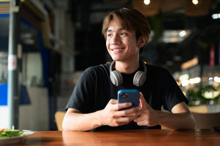 Portrait of a young Thai Asian man in casual style using mobile phone while sitting during day. Confident, relaxed, and authentic look suitable for various concepts and usesの写真素材