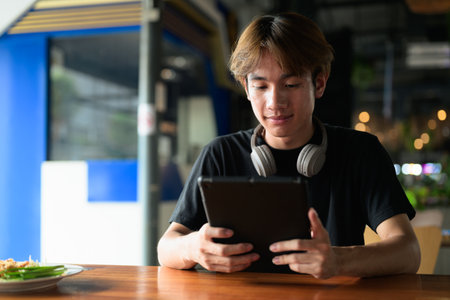 Young Thai Asian man using digital tablet computer during day while sitting in restaurant. Confident, relaxed, and authentic look suitable for various concepts and usesの写真素材