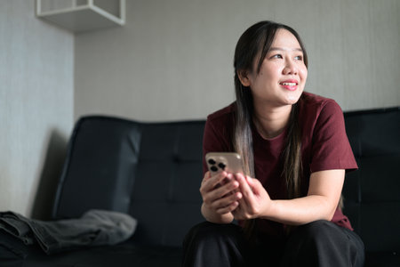 Young Thai woman sitting on sofa at home using smartphone. Modern lifestyle portrait showing technology, relaxation, and everyday communication.の写真素材