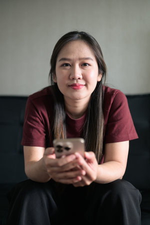 Young Thai woman sitting on sofa at home using smartphone. Modern lifestyle portrait showing technology, relaxation, and everyday communication.の写真素材