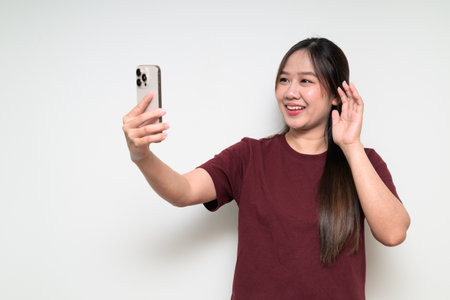 Studio portrait of young cute Asian woman using mobile phone against white background wearing casual t-shirt having video call meeting or doing selfie pictureの写真素材