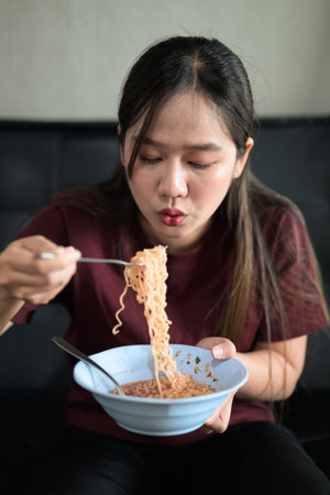 Portrait of Asian woman sitting at home in sofa eating instant noodles from bowl during day for lunchの写真素材
