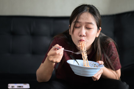 Portrait of Asian woman sitting at home in sofa eating instant noodles from bowl during day for lunchの写真素材