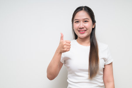 Studio portrait of young cute Asian woman doing thumbs up gesture against plain background wearing casual white t-shirtの写真素材