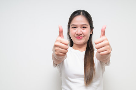 Studio portrait of young cute Asian woman doing thumbs up gesture against plain background wearing casual white t-shirtの写真素材