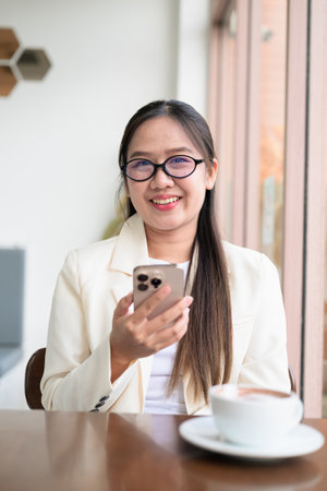 Young Asian businesswoman sitting in coffee shop restaurant wearing suit and eyeglasses while using smartphoneの写真素材