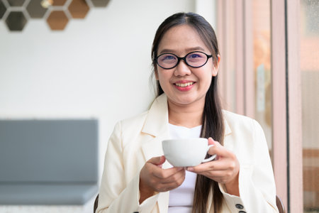 Young Asian businesswoman sitting in coffee shop restaurant wearing suit and eyeglasses drinking coffeeの写真素材