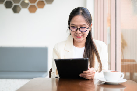 Young Asian businesswoman sitting in coffee shop restaurant wearing suit and eyeglasses while remotely working with digital tablet computerの写真素材