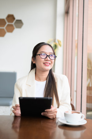 Young Asian businesswoman sitting in coffee shop restaurant wearing suit and eyeglasses while remotely working with digital tablet computerの写真素材